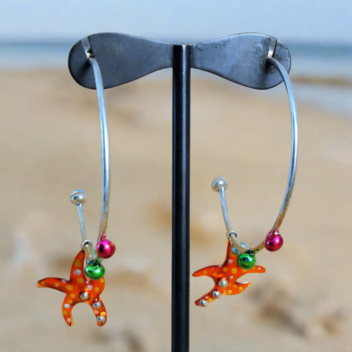 Handmade sterling silver hoop earrings with a vibrant orange starfish enamel design, displayed on a stand with a sandy background.
