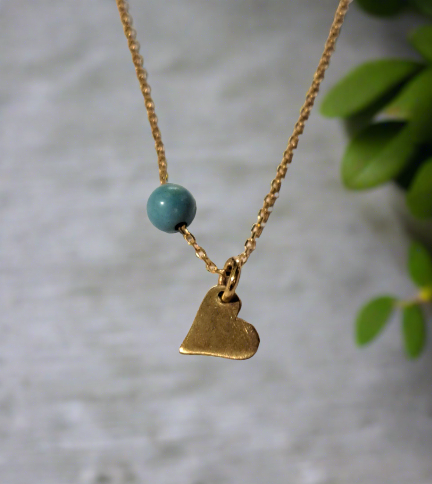 A 14K gold necklace with a small heart-shaped pendant and a turquoise bead on a chain, displayed against a neutral background with green foliage in the peripheral.