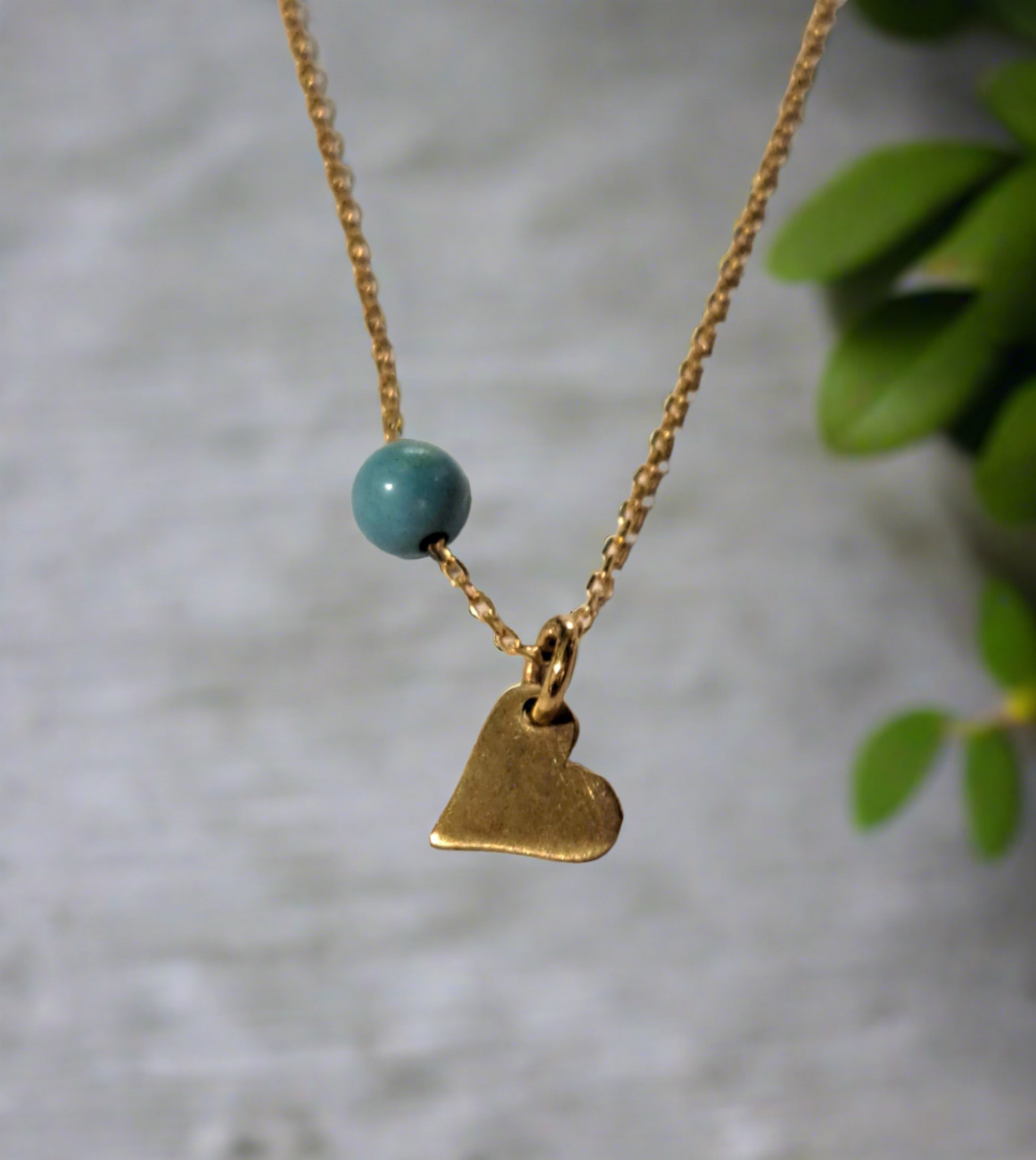 A 14K gold necklace with a small heart-shaped pendant and a turquoise bead on a chain, displayed against a neutral background with green foliage in the peripheral.