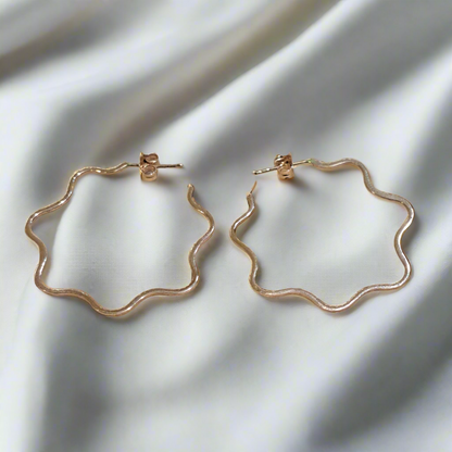 A pair of 14K red gold hoop earrings with a wavy motion design, displayed on a white silky surface.
