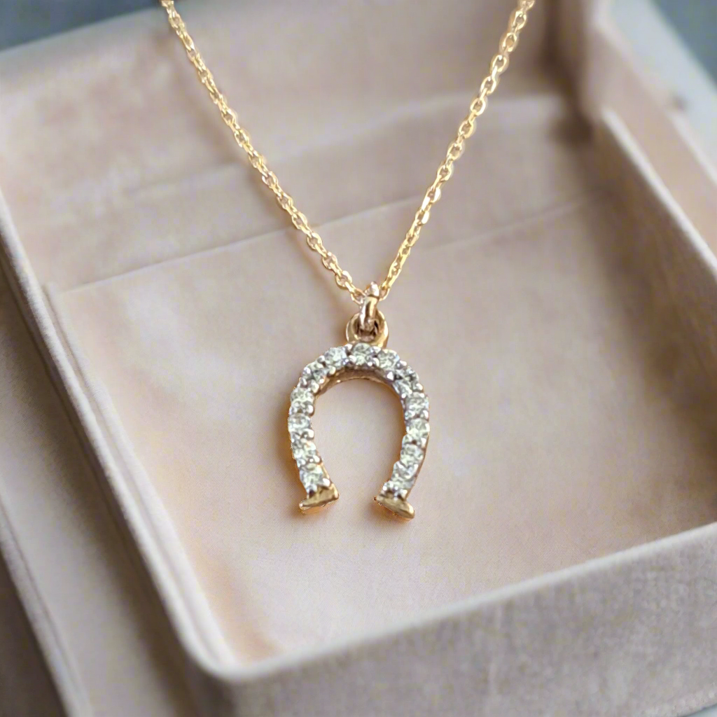 A 9K gold necklace with a horse shoe pendant embellished with sparkling zirconia, presented on a beige box.