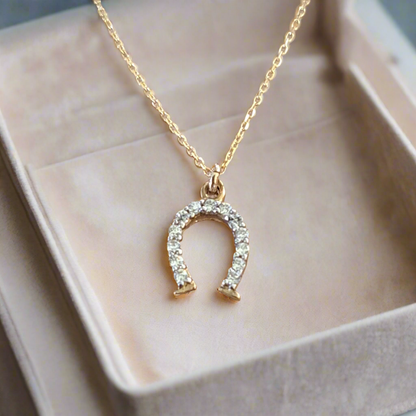 A 9K gold necklace with a horse shoe pendant embellished with sparkling zirconia, presented on a beige box.