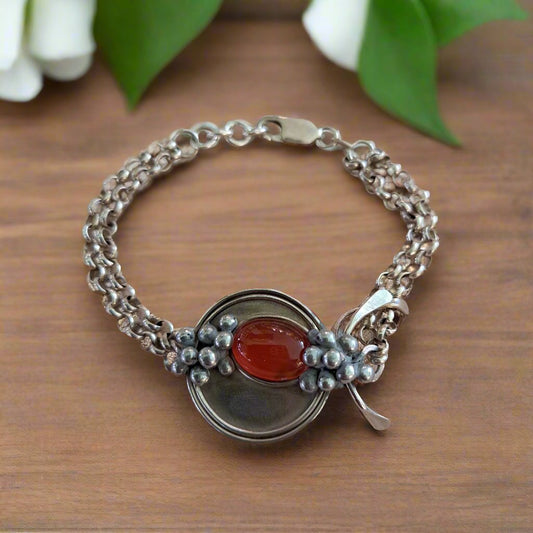 A handcrafted sterling silver bracelet with a central oval cornelian cabochon, surrounded by steling silver chains, displayed on a wooden surface with a floral accent in the background.