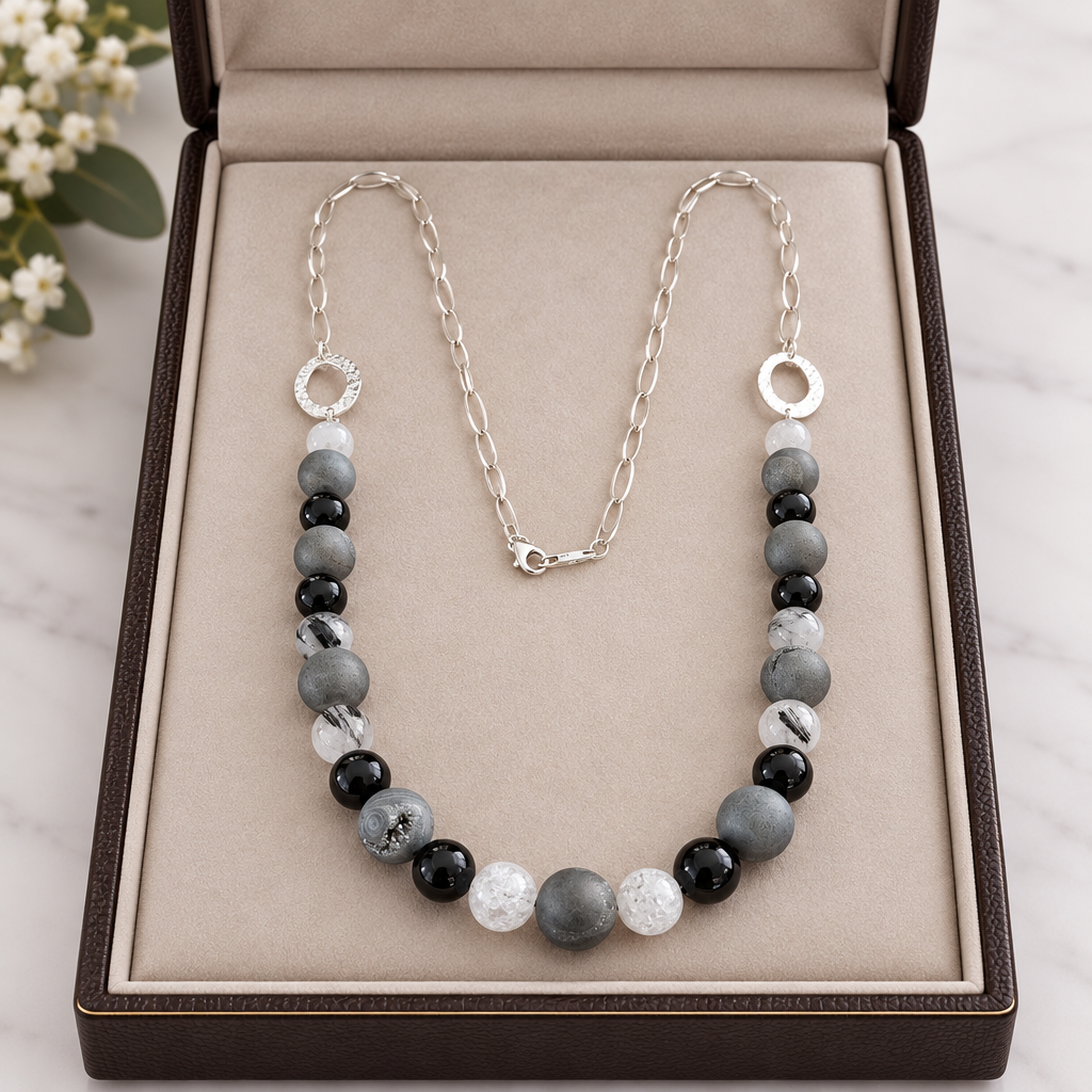 A beaded necklace with alternating black, gray, and white agates featuring a silver chain with a textured silver elements  displayed in a jewelry box