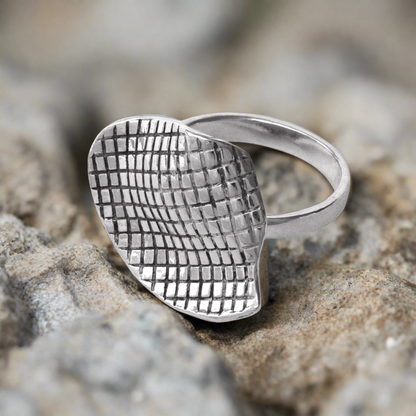 A silver ring with a geometric grid pattern on the top half, placed on a textured rocky surface.