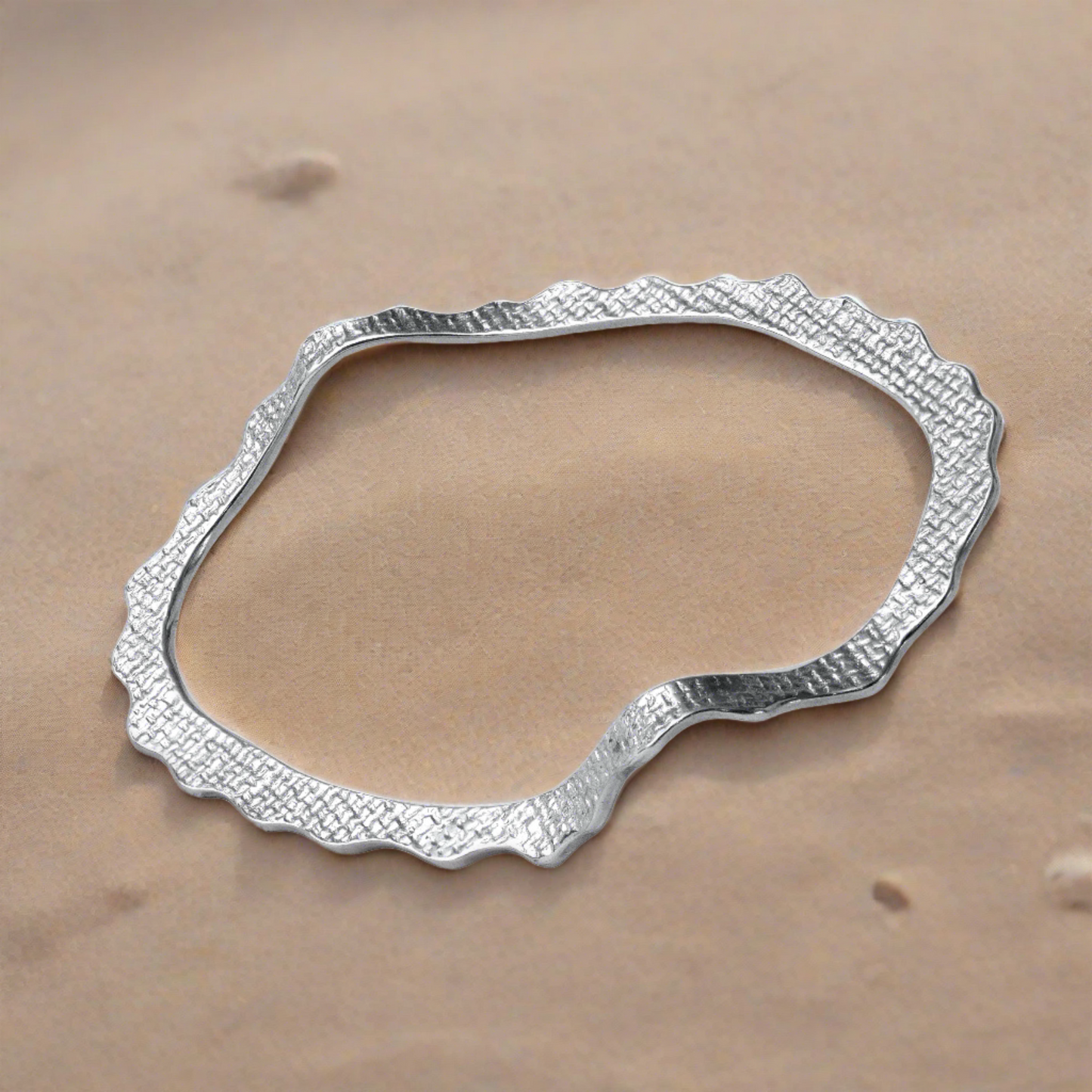 A sterling silver bracelet with a tiled texture, designed in a wave-like shape, displayed at the sand