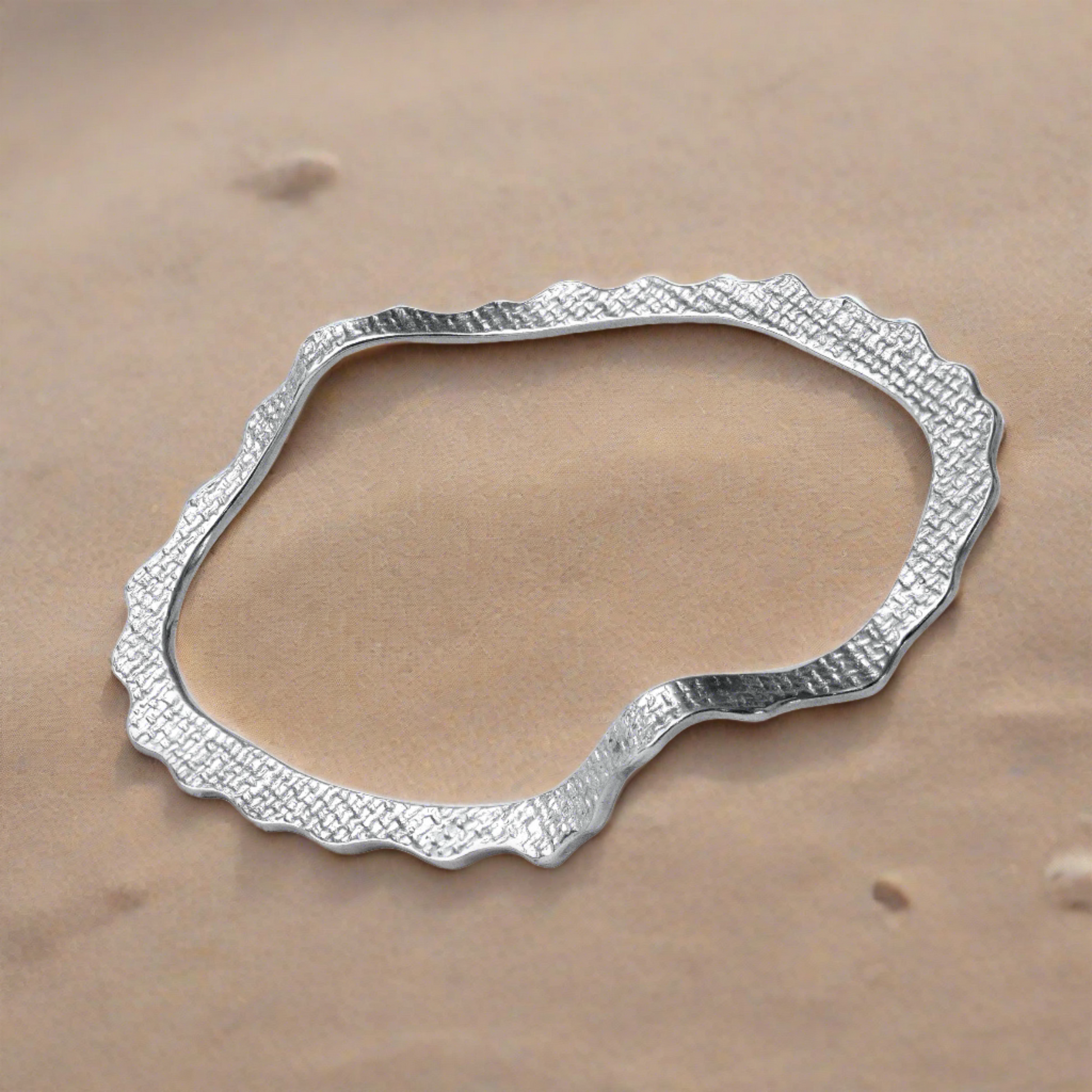 A sterling silver bracelet with a tiled texture, designed in a wave-like shape, displayed at the sand