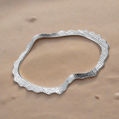 A sterling silver bracelet with a tiled texture, designed in a wave-like shape, displayed at the sand