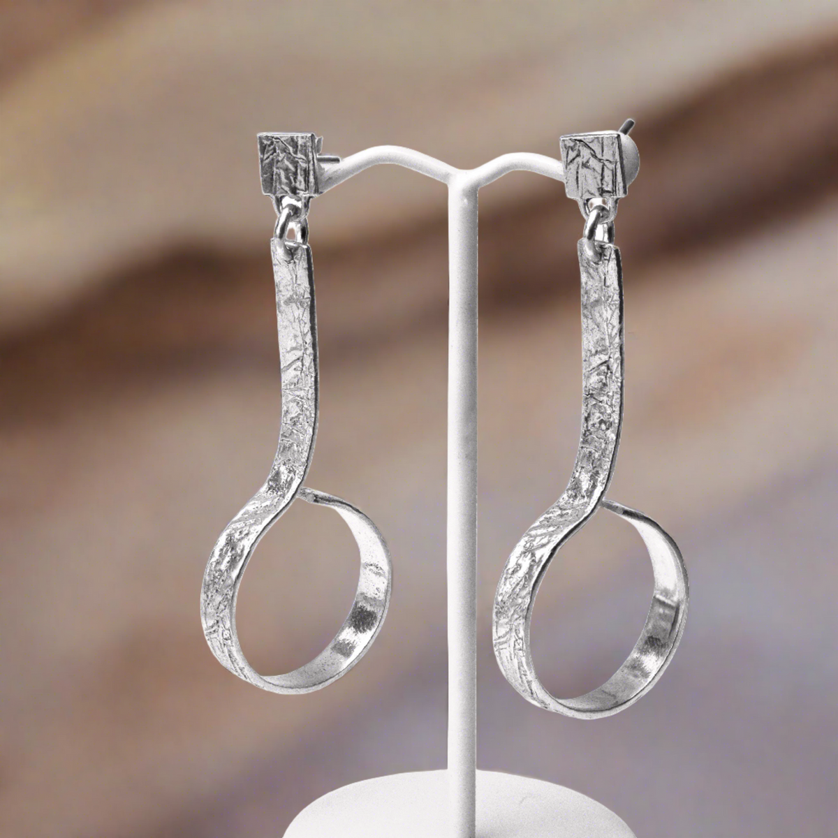 A pair of silver hoop earrings with intricate engravings, displayed on a white stand against a textured background.