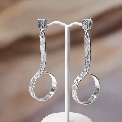 A pair of silver hoop earrings with intricate engravings, displayed on a white stand against a textured background.