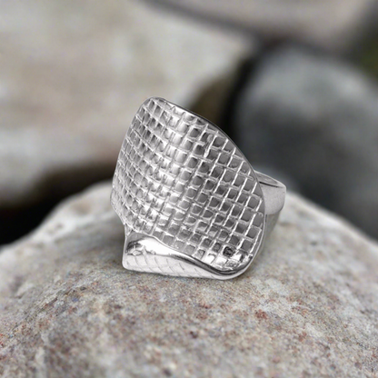 A sterling silver ring with a textured geometric pattern, placed on a rocky surface.