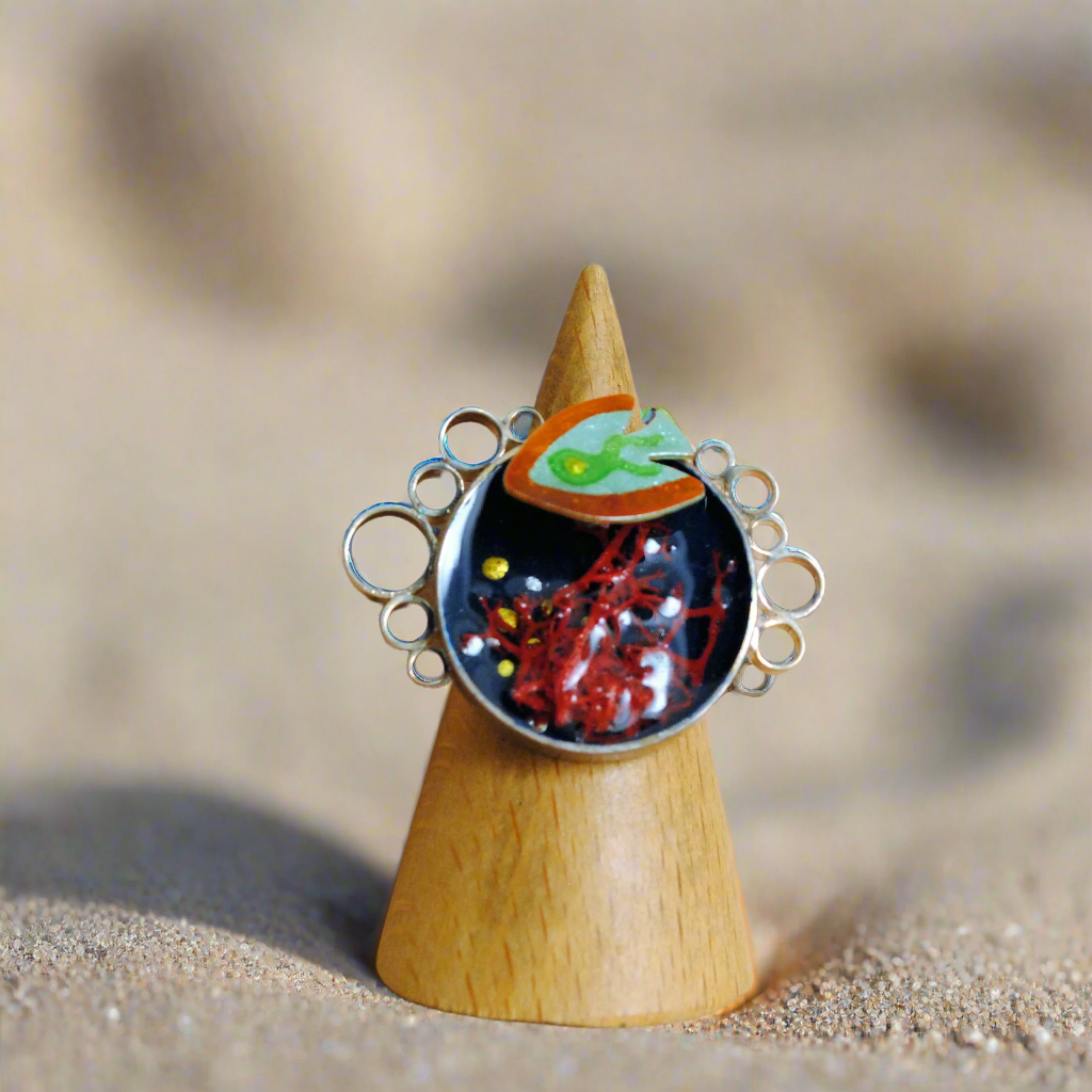 Silver ring with  a sea bed design with a fish, corals and bubbles painted by hand with  enamel on a wooden stand against a sandy background