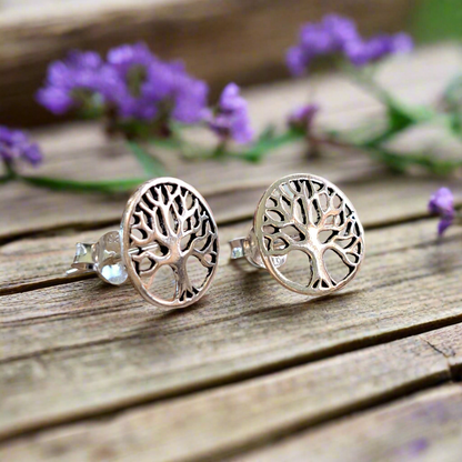 A pair of sterling silver tree of life stud earrings placed on a wooden surface with purple flowers in the background.
