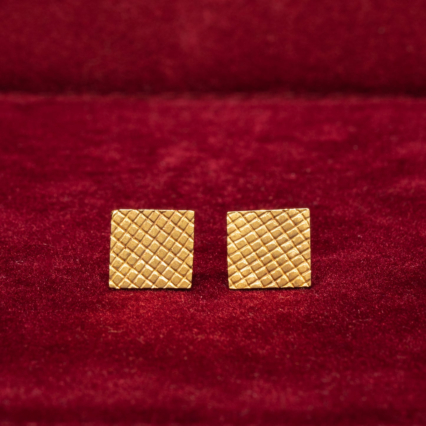 A pair of 14K gold square-textured stud earrings placed on a red surface.