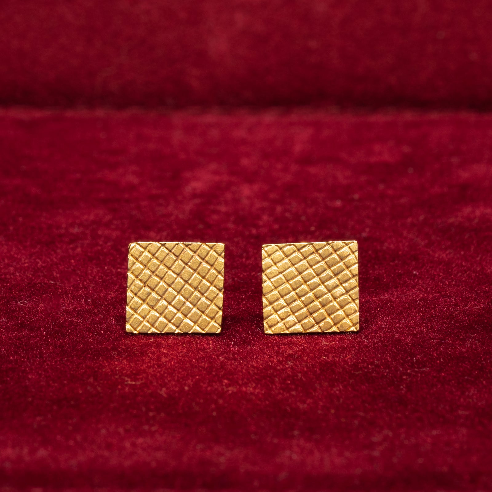A pair of 14K gold square-textured stud earrings placed on a red surface.