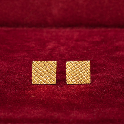 A pair of 14K gold square-textured stud earrings placed on a red surface.