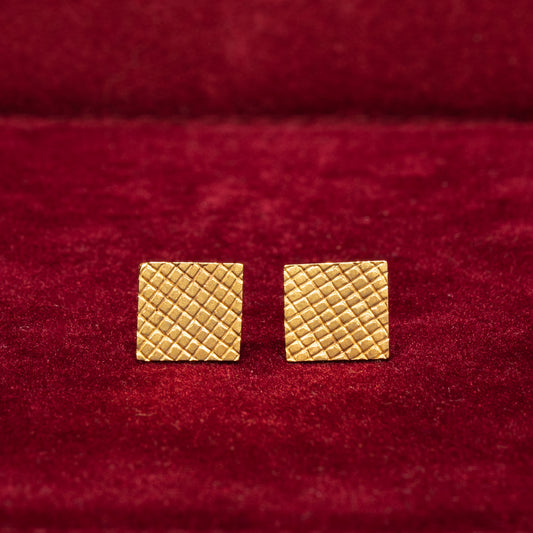 A pair of 14K gold square-textured stud earrings placed on a red surface.