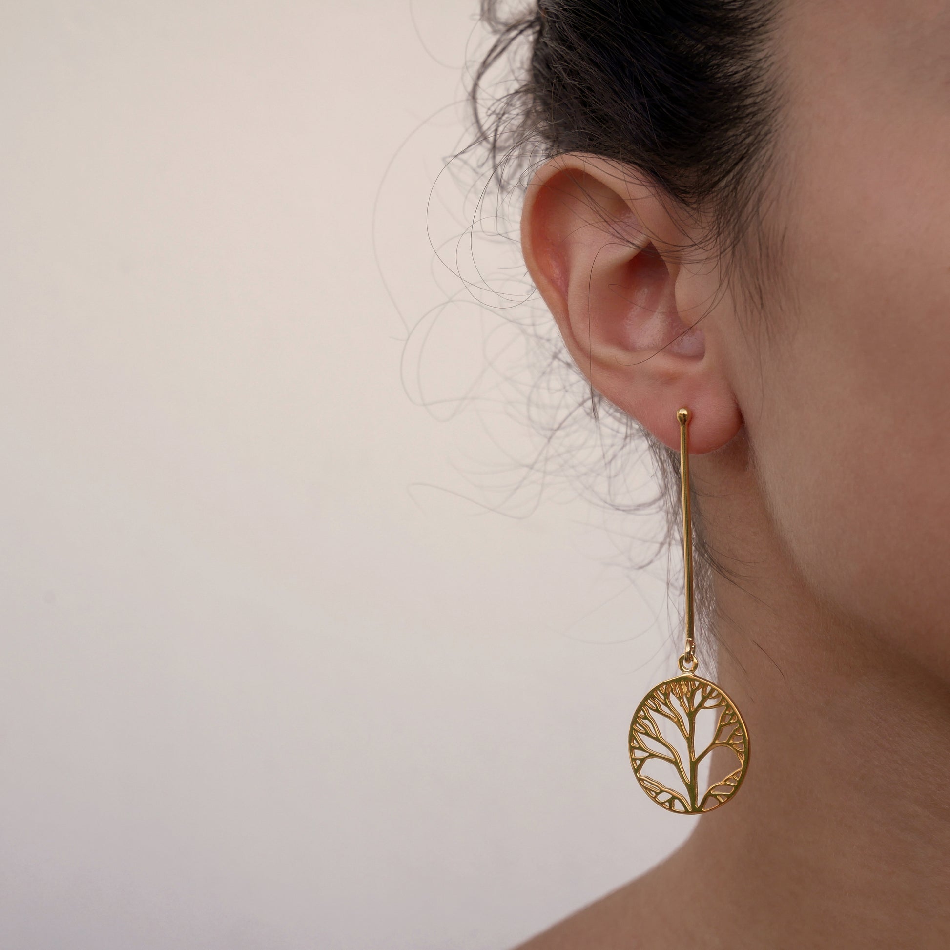 A woman's face wearing a long gold plated silver earring . In a verical massive wire hangs a circle with the design of a tree.  The tree is delicate and beautiful.